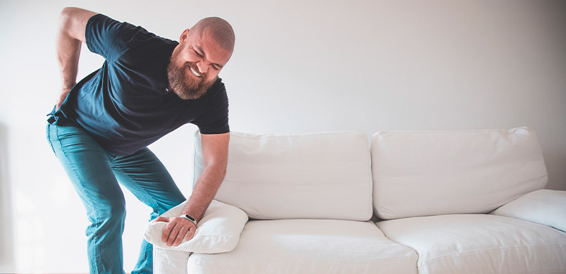 A man is leaning over a couch, possibly searching for an item or adjusting something on it. Back injury, back pain. 