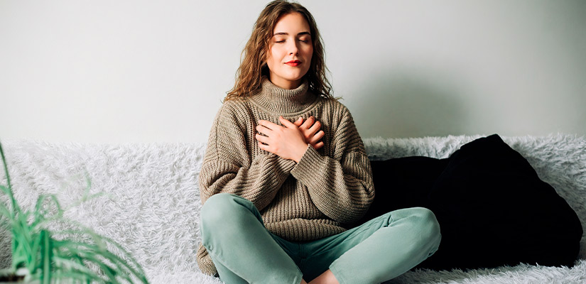 A woman seated on a couch, hands on her chest doing breathing exercises, illustrating the connection between holiday stress and physical discomfort.