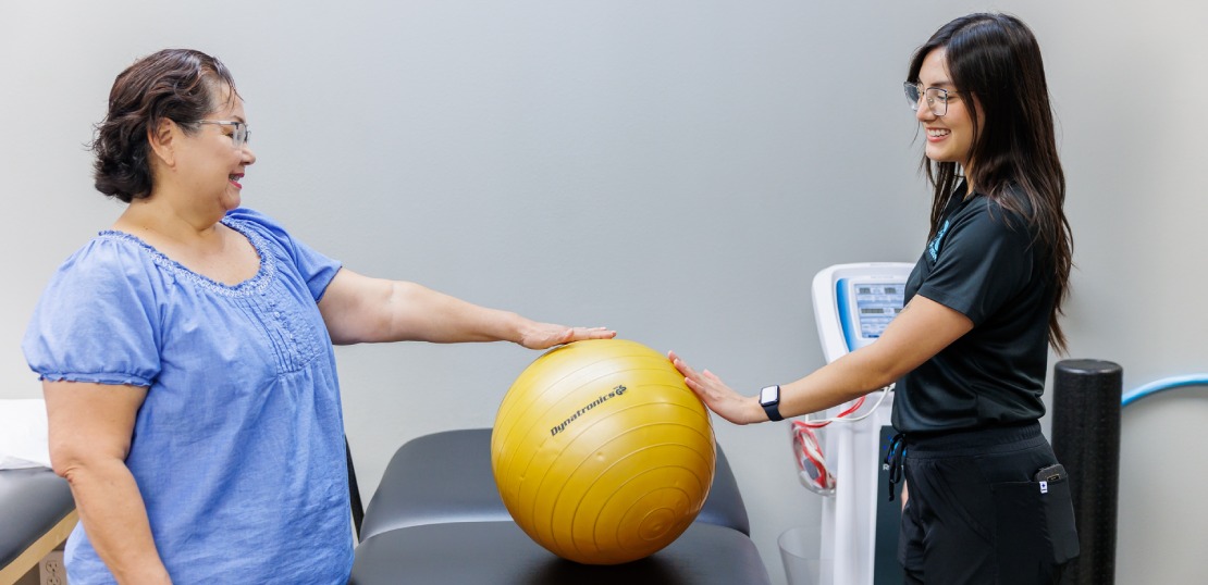 A woman holds an exercise ball while a physical therapist beside her, demonstrating functional therapy techniques.
